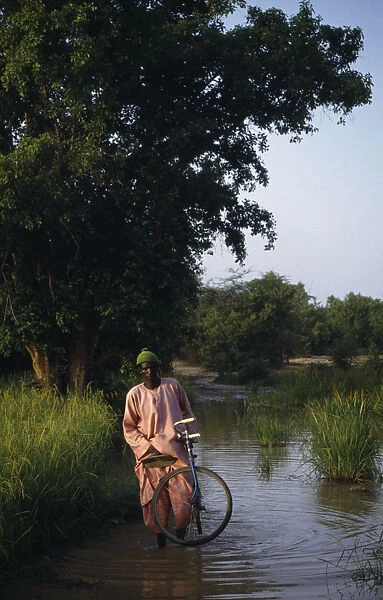 20075224. GAMBIA People Men Man with bike wading through water