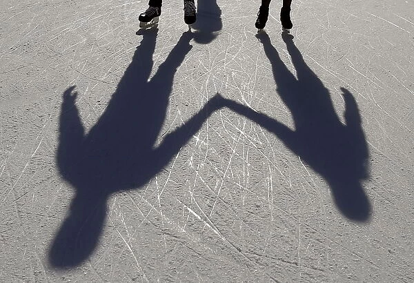 A couple casts shadows on the ice as they hold hands and skate on an outdoor rink