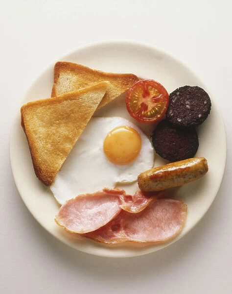 Plate of full English breakfast, fried egg, slices of bacon, sausage, toast, fried tomato and black pudding