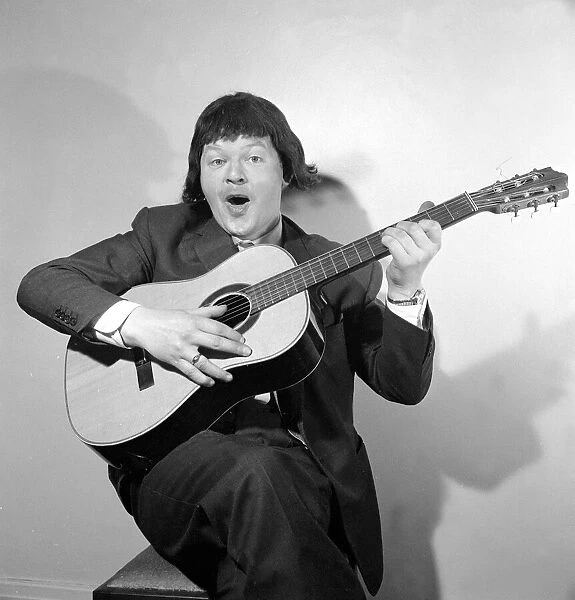 Comedian Benny Hill pictured at home wearing a wig and playing the guitar May 1958