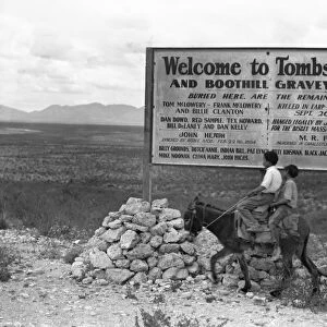ARIZONA: TOMBSTONE, 1937. Two boys riding a donkey, in front of a sign entering Tombstone, Arizona and Boothill Graveyard. Photograph by Dorothea Lange, 1937
