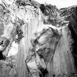 Excavators at work along a steep wall of a marble quarry in the Apuan Alps, Tuscany. (b / w photo)
