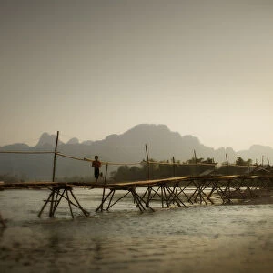 Boy Running Across A Wooden Bridge On A River With Mountains In The Background; Vang Vieng Laos