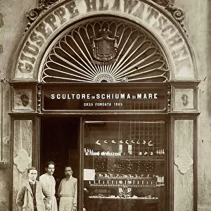 Sign and window of a shop with sepiolite sculpture. At the entrance door: two people in work clothes together with a man